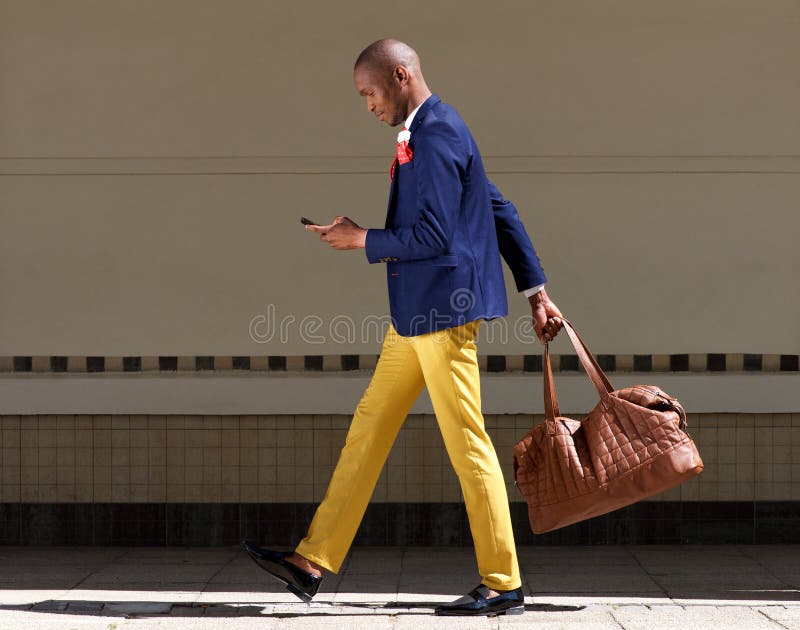 Full length side portrait of young african businessman using mobile phone and walking outdoors on the road. Full length side portrait of young african businessman using mobile phone and walking outdoors on the road