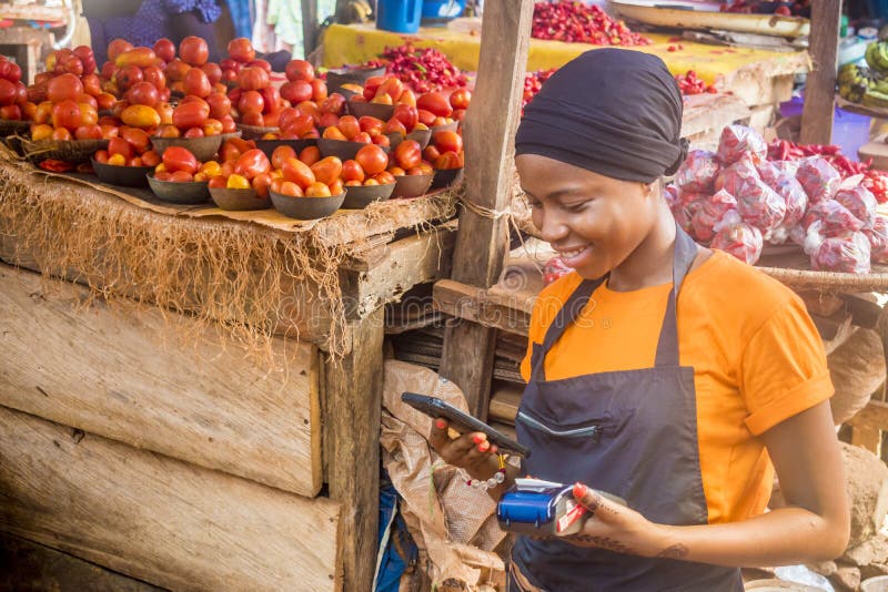 african woman selling in a local african market holding a mobile point of sale system and using her mobile phone