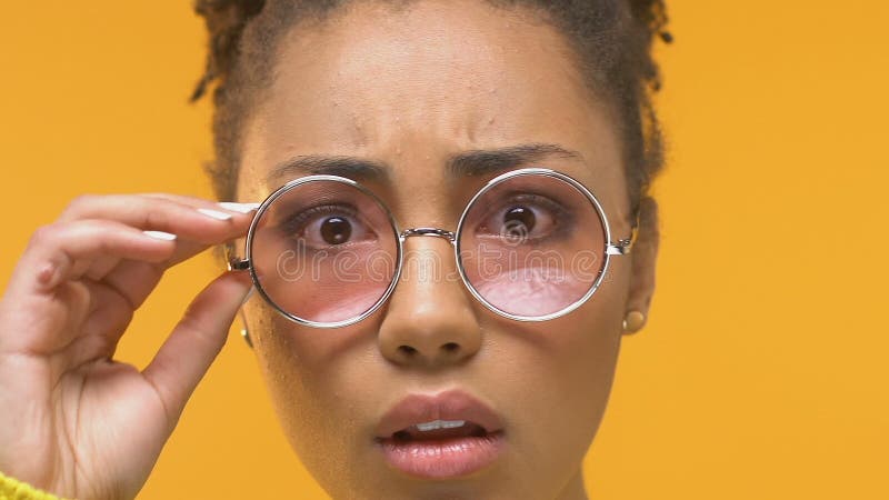 Young african woman taking off eyeglasses looking shocked on camera, dermatology