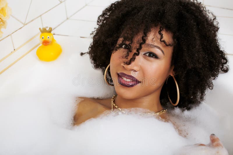 Young afro-american teen girl laying in bath with foam, wearing swag jewelry flawless, making selfie