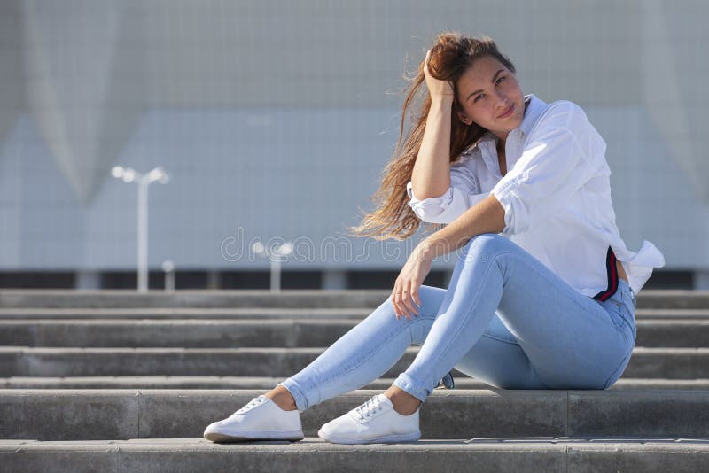 Young American Woman In A White T Shirt In Jeans In White Trendy Sneakers Relaxes Sitting On The Steps Cute European Girl Model Stock Photo Image Of Caucasian Building
