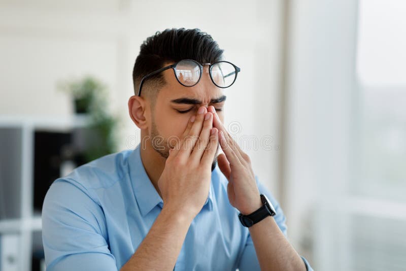 Young Arab office worker in glasses rubbing tired eyes, exhausted from overworking at modern office, copy space