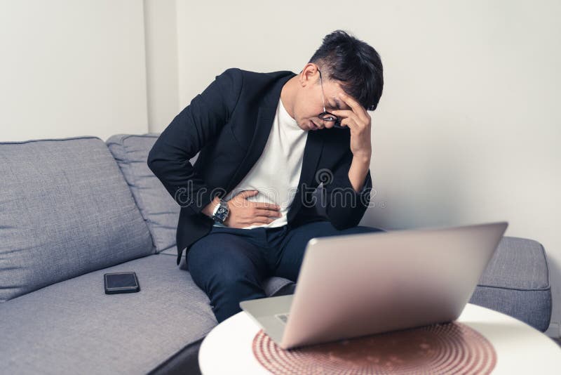 Young Asian business man sitting on sofa and working from home. Man feeling stress and suffering from headache and stomachache.