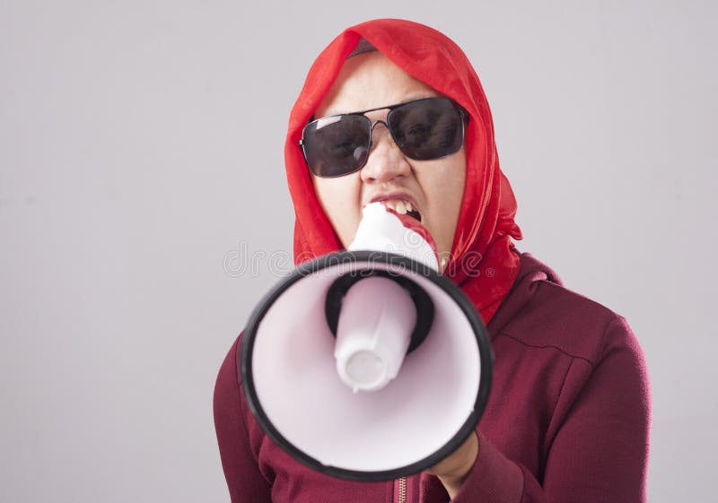 Young Asian businesswoman wearing red suit and hijab shouting with megaphone, angry expression. Close up body portrait. Young Asian businesswoman wearing red suit and hijab shouting with megaphone, angry expression. Close up body portrait