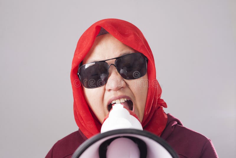 Young Asian businesswoman wearing red suit and hijab shouting with megaphone, angry expression. Close up body portrait. Young Asian businesswoman wearing red suit and hijab shouting with megaphone, angry expression. Close up body portrait