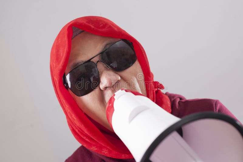 Young Asian businesswoman wearing red suit and hijab shouting with megaphone, angry expression. Close up body portrait. Young Asian businesswoman wearing red suit and hijab shouting with megaphone, angry expression. Close up body portrait