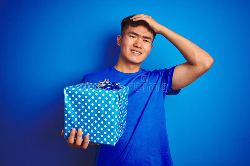 Young asian chinese man holding birthday gift standing over isolated blue background stressed with hand on head, shocked with stock image
