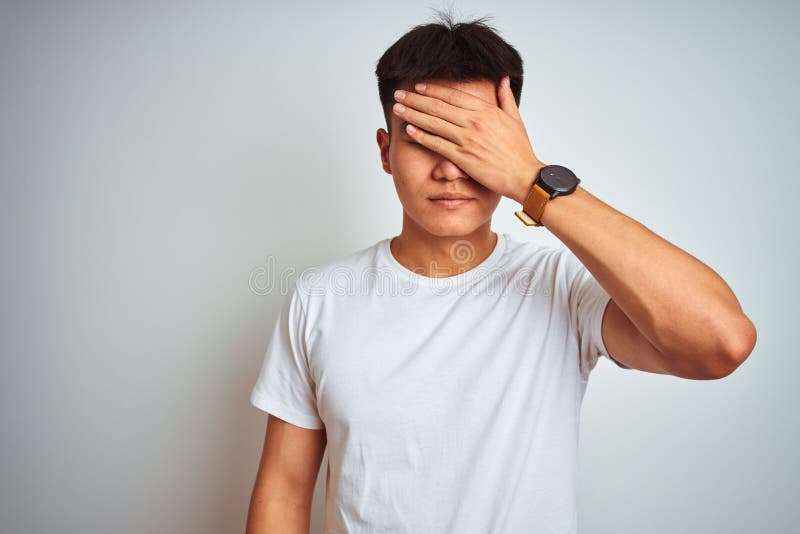 Young asian chinese man wearing t-shirt standing over isolated white background covering eyes with hand, looking serious and sad