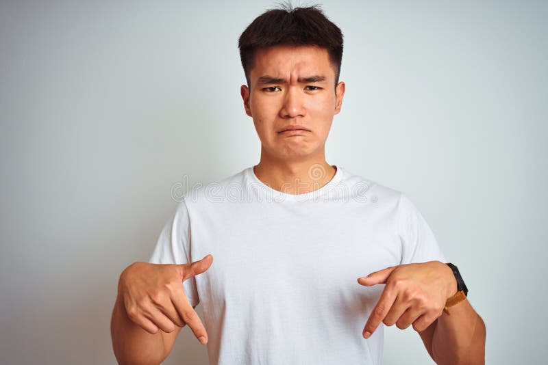 Young asian chinese man wearing t-shirt standing over isolated white background Pointing down looking sad and upset, indicating