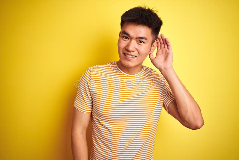 Young asian chinese man wearing t-shirt standing over isolated yellow background smiling with hand over ear listening an hearing royalty free stock photography