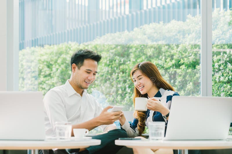 Young Asian couple, coworkers, or business partners have fun using smartphone together, with laptop computer at coffee shop. Information technology, cafe lifestyle, or romantic relationship concept