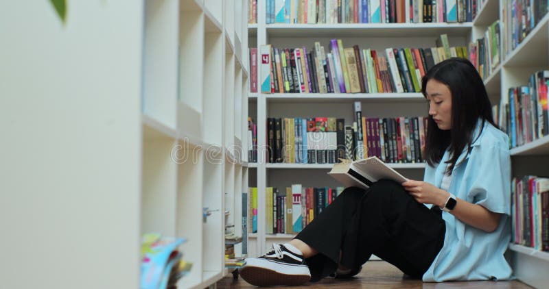 Young asian girl student reading book, sitting among stack of books on library floor, side view.