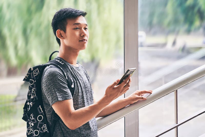 Young asian male student with smartphone in hand wearing t-shirt and backpack standing on university stairs and looks stock photo Young asian male student with smartphone in hand wearing t-shirt and backpack standing on university stairs and looks stock photo