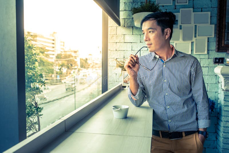 Young Asian man hand hold eye glasses looking out of window, Casual professional entrepreneur working-in out of office in cafe or