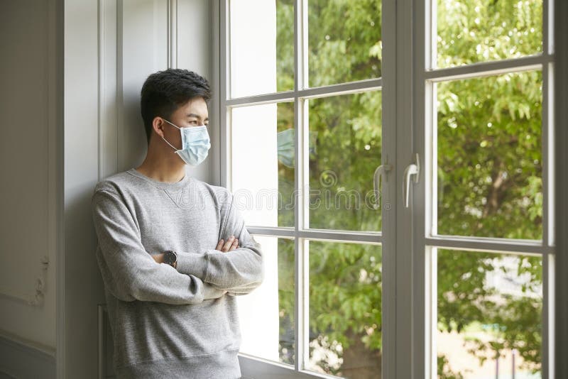 Young asian man wearing mask standing by winow at home