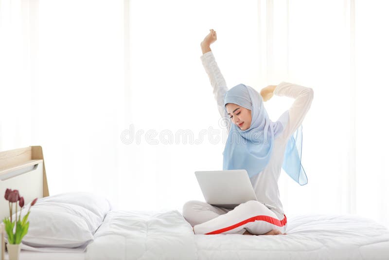 Young asian muslim woman using laptop sitting on bed in bedroom.