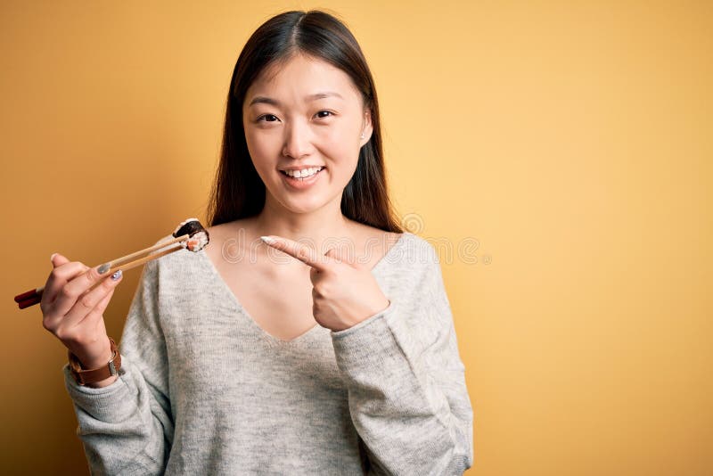 Young asian woman eating japanese food, holding salmon and rice maki sushi using chopsticks very happy pointing with hand and royalty free stock images
