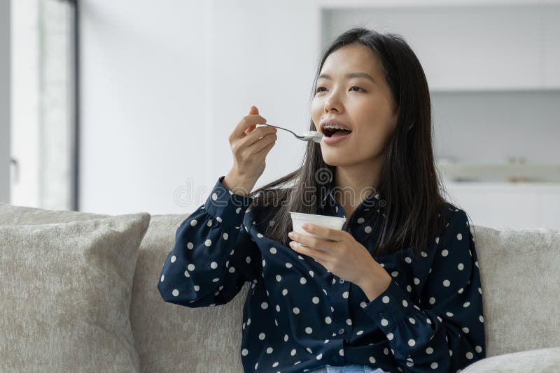 Young Asian Woman Eats Fresh Yogurt For Breakfast Sitting On The Couch