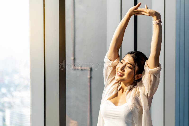 Young asian woman standing in front of big window, stretching arms and back, enjoying good morning time in office.