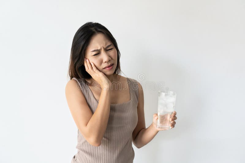 Young Asian woman suffering from toothache and hand holding glass of cold water with ice. Girl drinking cold drink, glass full of