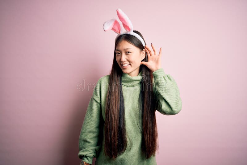 Young asian woman wearing cute easter bunny ears over pink background smiling with hand over ear listening an hearing to rumor or stock photo