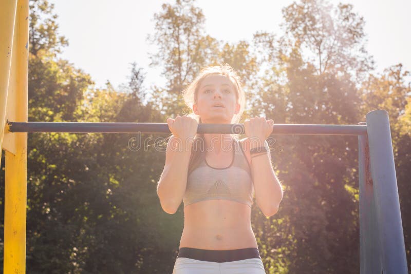 Young athletic fitness woman working out at outdoor gym doing pull ups at sunrise.