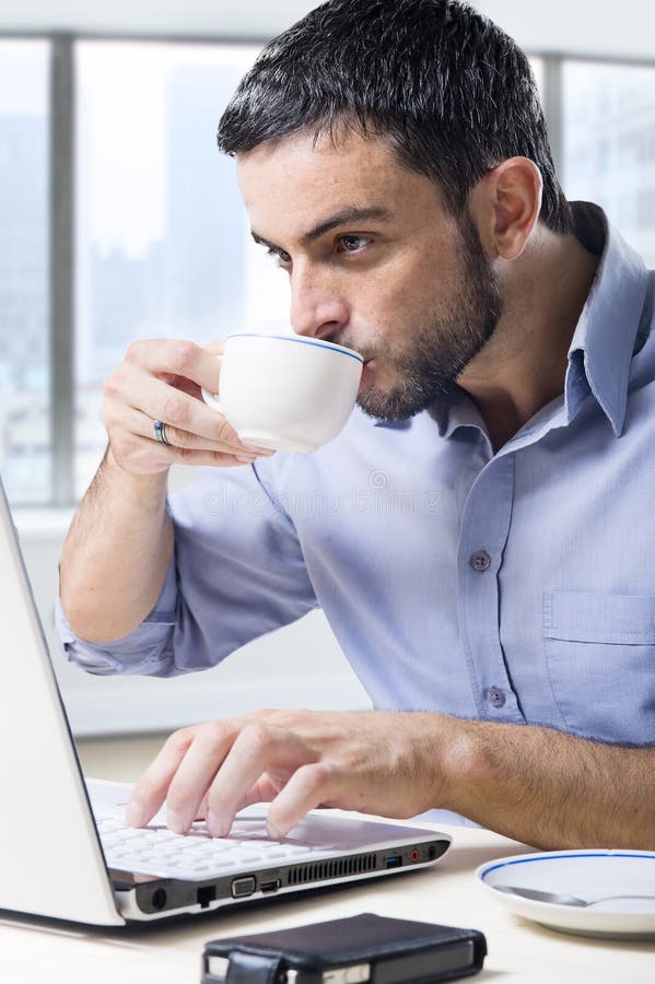 Young attractive businessman working on computer laptop drinking cup of coffee cup sitting at office desk royalty free stock photos