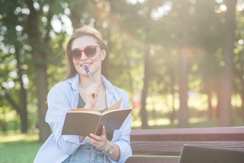 A young attractive Caucasian girl sitting on a bench in the park, holding a notepad and thinking. Freelancer, student. Education concept