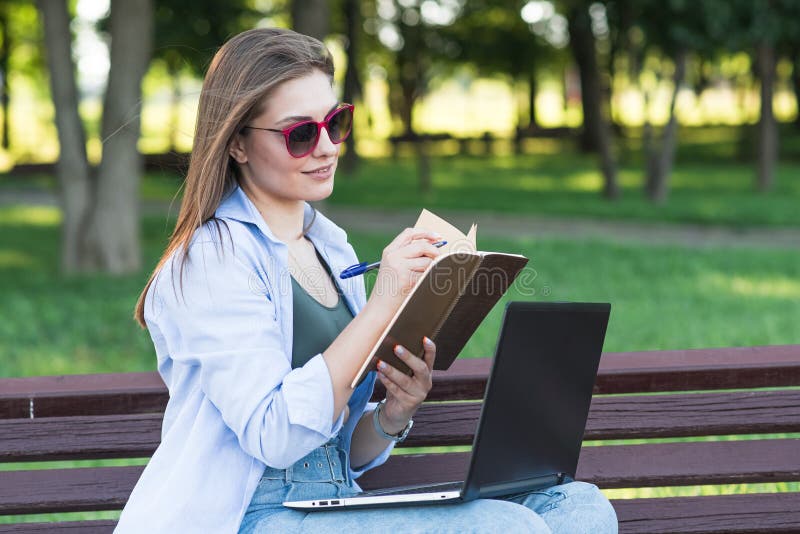 A young attractive Caucasian girl sitting on a bench in the park and writing in a notebook. Freelancer, student. Education concept