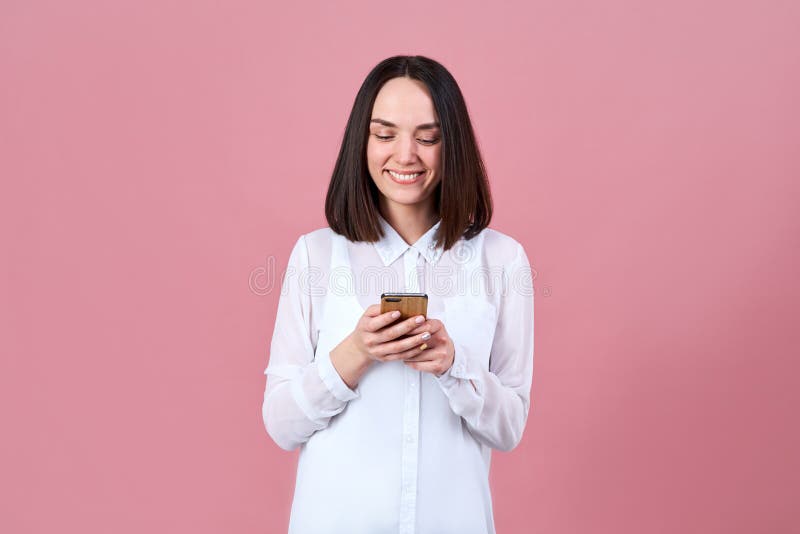 A young attractive friendly brunette woman in a white shirt smiles widely with her teeth and writes a message on her smartphone. Studio portrait on pink isolated background