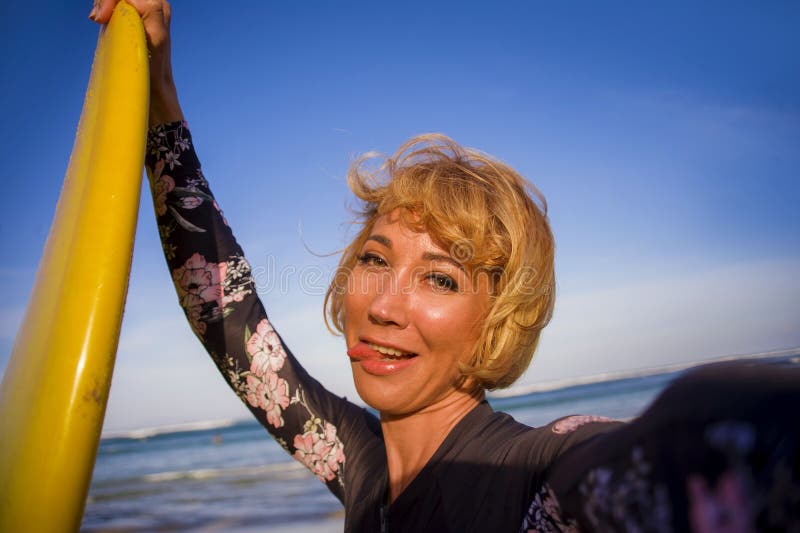 Young attractive and happy blonde surfer woman in swimsuit holding surf board in the beach taking self portrait selfie picture smi royalty free stock image Young attractive and happy blonde surfer woman in swimsuit holding surf board in the beach taking self portrait selfie picture smi royalty free stock image
