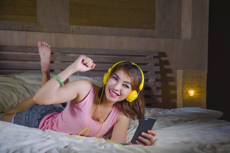 Young attractive and happy girl in bed with yellow headphones listening to music on internet with mobile phone dancing and singing