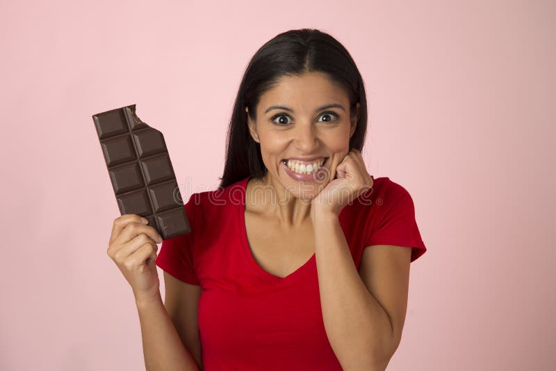 Young attractive and happy hispanic woman in red top smiling excited eating chocolate bar on pink background stock photography Young attractive and happy hispanic woman in red top smiling excited eating chocolate bar on pink background stock photography