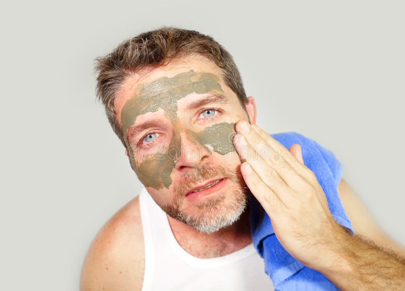 Young attractive and happy man with bathroom towel with green cream on his face applying facial mask in skin care beauty