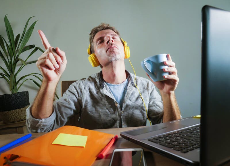 Young attractive and happy man with yellow headphones sitting at home office desk working with laptop computer having fun listenin