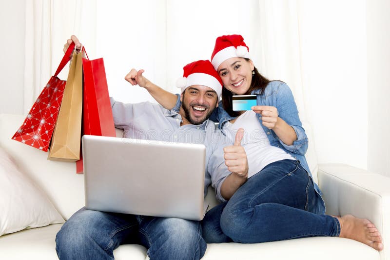 Young attractive Hispanic couple in love together on sofa couch at home with computer laptop, holding shopping bags purchasing online Christmas presents with credit card on internet. Young attractive Hispanic couple in love together on sofa couch at home with computer laptop, holding shopping bags purchasing online Christmas presents with credit card on internet