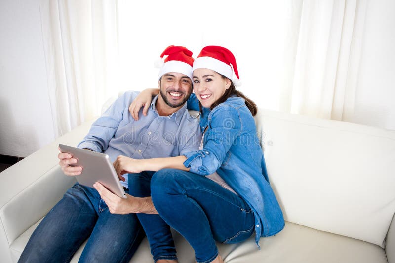 Young attractive Hispanic couple in love together on sofa couch wearing Santa hat with digital tablet computer smiling happy purchasing online Christmas presents on internet. Young attractive Hispanic couple in love together on sofa couch wearing Santa hat with digital tablet computer smiling happy purchasing online Christmas presents on internet