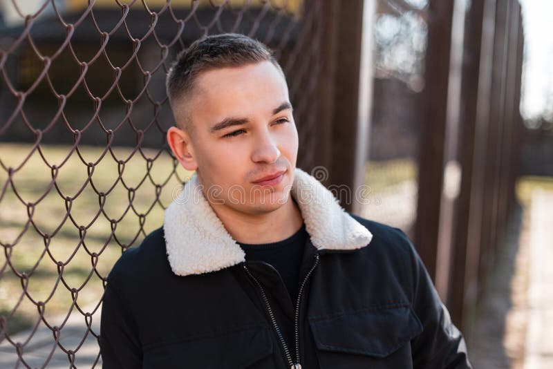 Young attractive man in a black stylish jacket with a white collar in a trendy T-shirt is rest standing near a metal vintage fence