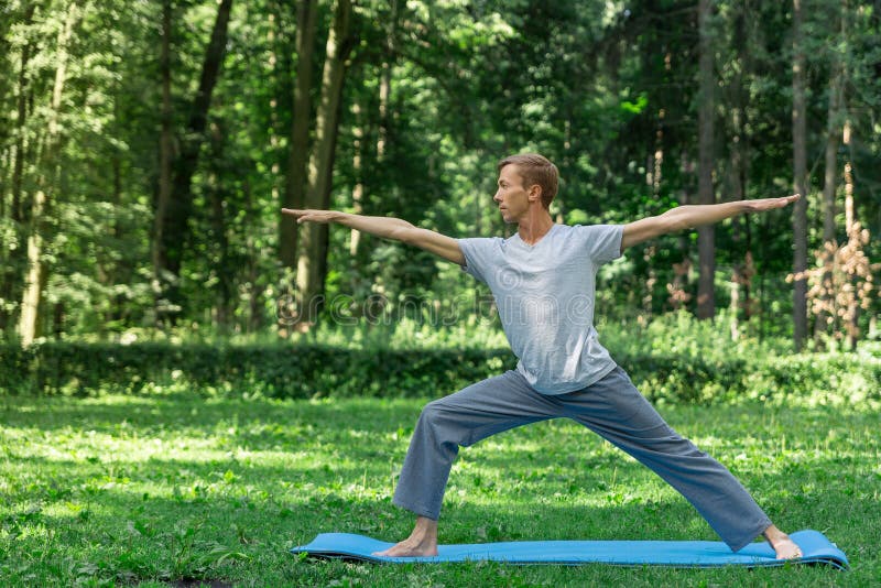 Young attractive man in a gray T-shirt and sweatpants doing yoga warrior pose in the park. He spread his legs wide, royalty free stock photo Young attractive man in a gray T-shirt and sweatpants doing yoga warrior pose in the park. He spread his legs wide, royalty free stock photo