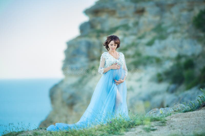 Young attractive pregnant lady woman wearing casual blue airy dress posing close to ocean sea cape mountains over sky background.