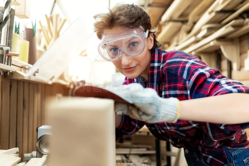 Young attractive 30-40 professional female carpenter looking grinding raw wood with sandpaper in carpentry diy workshop. Feminine