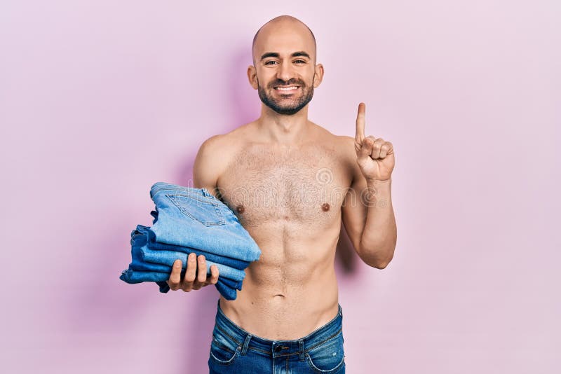Young bald man holding stack of folded jeans smiling with an idea or question pointing finger with happy face, number one royalty free stock image