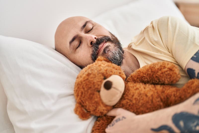 Young bald man hugging teddy bear lying on bed sleeping at bedroom royalty free stock photo