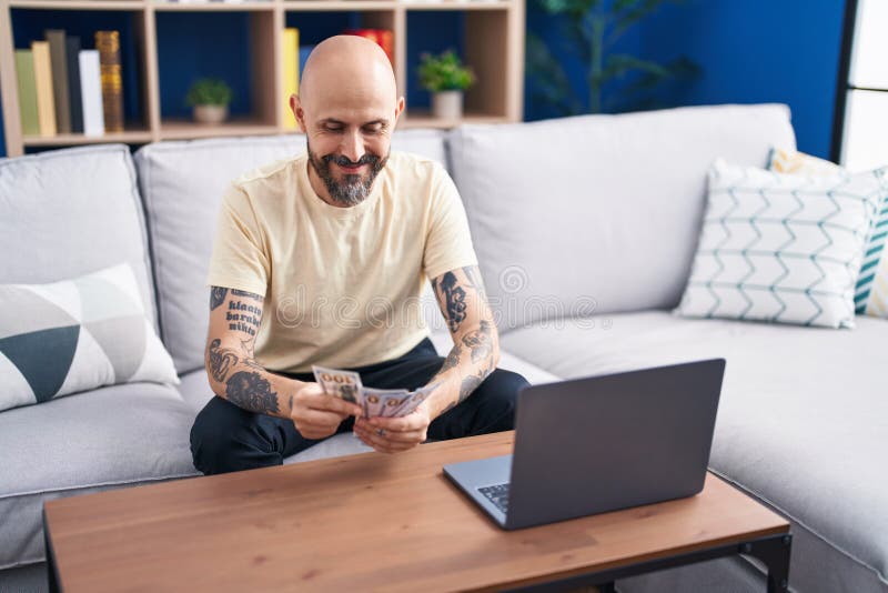 Young bald man using laptop counting dollars at home royalty free stock image