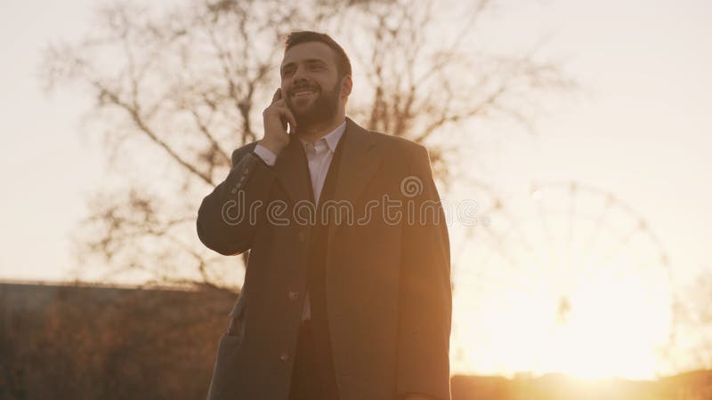 Young bearded business man in coat talking at smartphone and making deals on street with sunlight backlit in evening stock images Young bearded business man in coat talking at smartphone and making deals on street with sunlight backlit in evening stock images