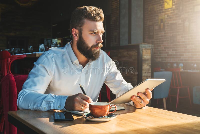 Young bearded businessman is sitting in cafe at table, holding tablet computer and writing in notebook.