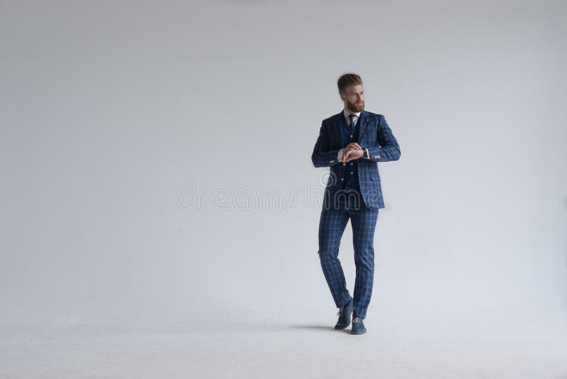 Full length shot of young bearded stylish businessman leader indoors dressed in three-piece suit checking the time on his watch.