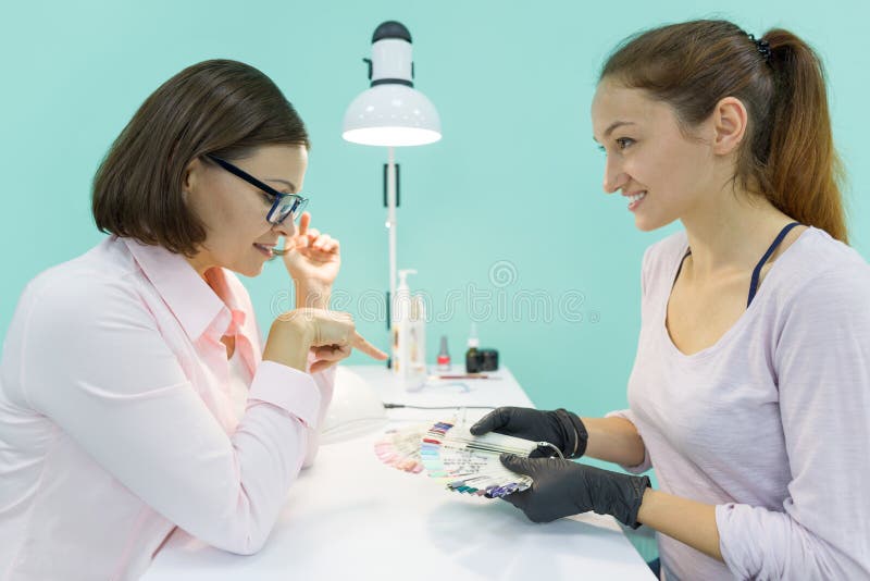 Young beautician girl manicurist talks to a female client, does manicure, chooses the color of nail polish, communicates and smile