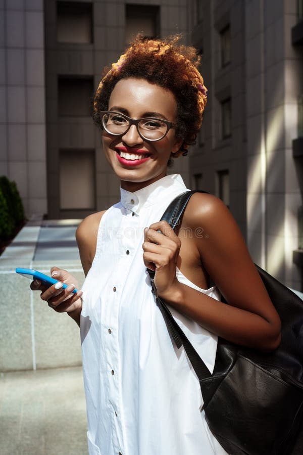 Young beautiful african girl smiling holding phone walking down city.