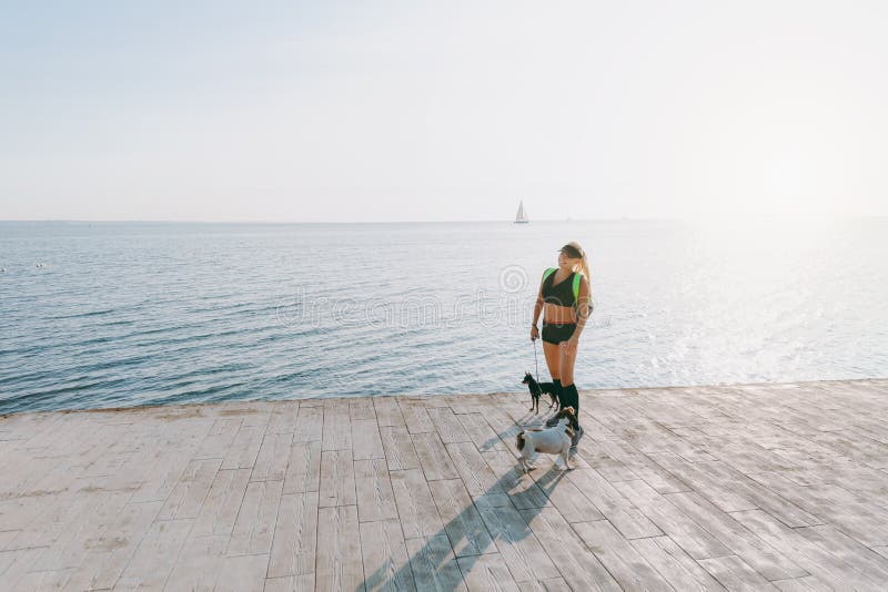 Young beautiful athletic girl with long blond hair in black clothes and two of her dogs doing sports at sunrise by the sea stock photos Young beautiful athletic girl with long blond hair in black clothes and two of her dogs doing sports at sunrise by the sea stock photos